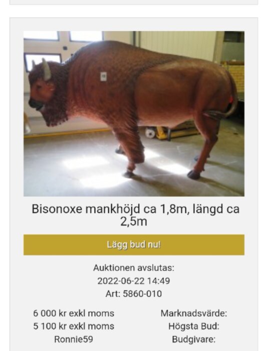 En konstgjord bisonoxe med mankhöjd ca 1,8 m och längd ca 2,5 m står på ett golv med en gul dörr och en gaffelvagn i bakgrunden. Auktionstext under bilden.