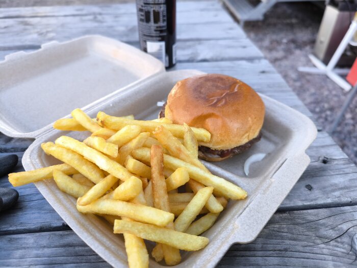 Hamburgare och pommes frites i en brun engångsbehållare på ett slitet träbord utomhus med en dryckesburk i bakgrunden.