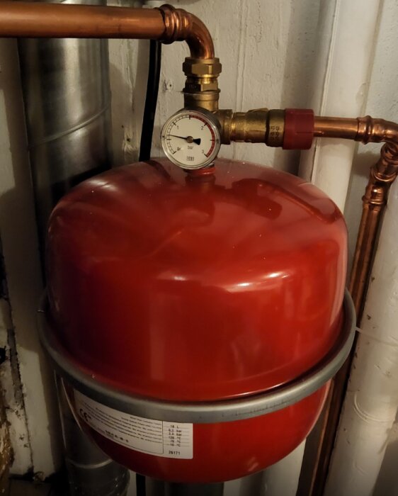 Rött expansionskärl med tryckmätare monterad upptill, visar tryck i bar, kopplad till kopparrör i ett utrymme med flera rörinstallationer.