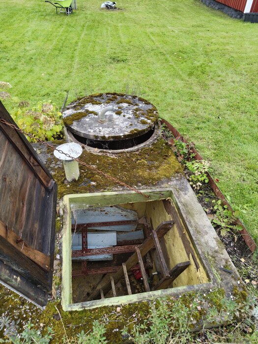 Gammalt pumprum med gjuten cistern ovanpå med mossa, inbäddat i gräsmatta. Öppen lucka visar stege och inre rummet.