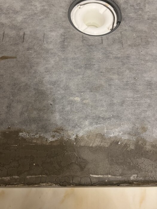 Sprickor i golv med någon slags beläggning eller kitt runt ett avloppshål, relaterat till pågående byggarbeten och härdning av material.