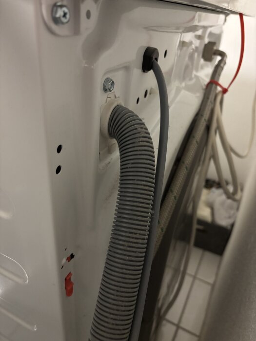 Grå avloppsslang ansluten till baksidan av en tvättmaskin, med synliga fästen och skruvar.