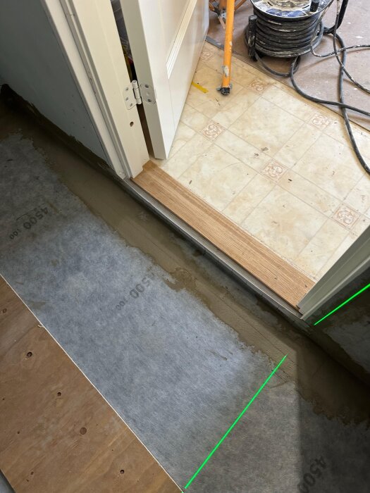 Tröskel vid en dörröppning, med golvmaterial och en grön laserlinje synlig på en underliggande yta.