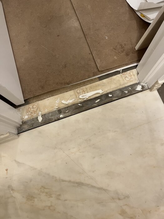 Tröskel mellan två rum, med gammalt kakel och plastgolv under. Tätskikt synligt och ojämnt underlag.
