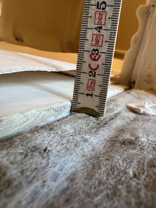 Mätsticka visar höjdskillnad mellan golvskikt intill en tröskel; mätsticka står på gammalt köksgolv nära ett modernt tätskikt i en byggprojektkontext.