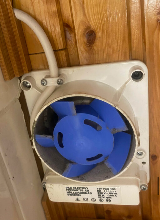 Blå paxfläkt installerad på trångt utrymme mot träpanel, visar platsbrist mellan fläkt och vägg.