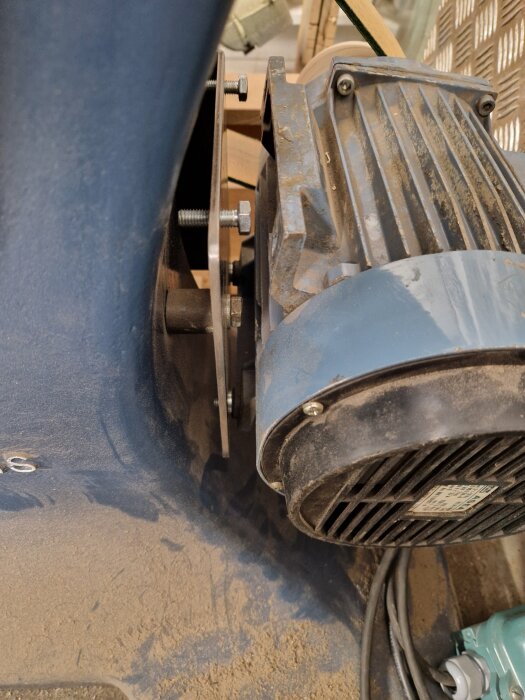 Närbild av motorfäste på bandsåg med monterad motor och sfäriska brickor, damm synligt på ytorna. Flatrem och drivhjul delvis i fokus.