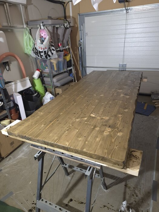 Träplanka under behandling i ett garage, färgad i ljusbrunt. Bakgrunden visar verktyg och garageutrustning.