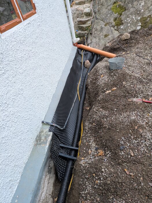 Knölig platonmatta och dräneringsrör vid en stenig husgrund, med stuprör och grus. En hink står nära grävområdet.