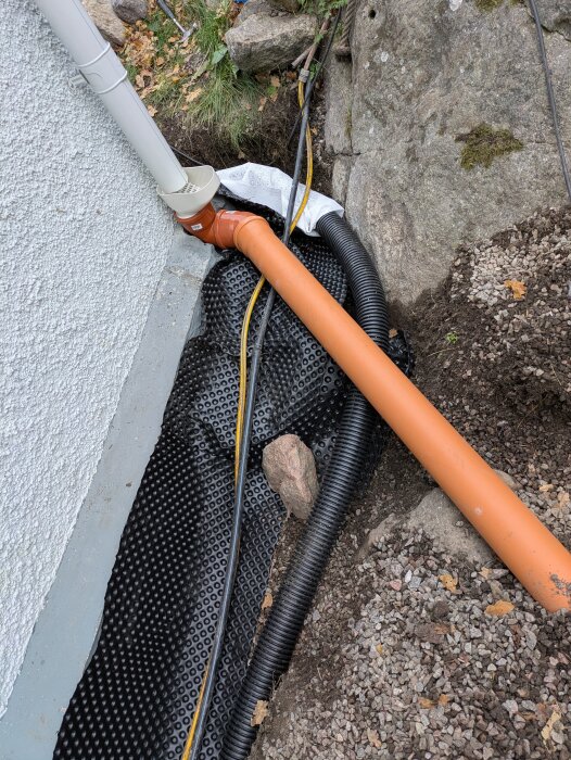 Knölig platonmatta med dräneringsrör intill stor sten, stuprör med lövavskiljare och orange avloppsledning, omgiven av grus och stenar.