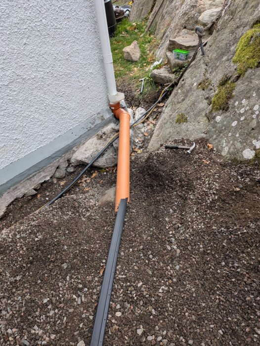 Dräneringsrör installerat vid husvägg, med orange avloppsrör och stuprör. Omgiven av grus och stenar, mot en bakgrund av mossklädd sten.