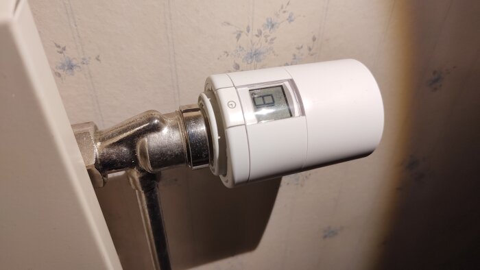 Danfoss ECO Bluetooth-regulator installerad på en radiator, viser en slät, vit design med en digital skärm mot en blommigt mönstrad tapet.