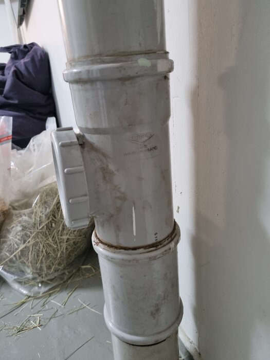 Grått avloppsrör med synliga skarvar, smuts och damm, stående mot en vit vägg.