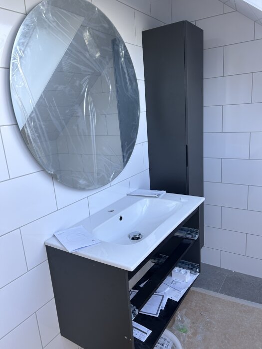 Badrum med vit kakelvägg, ovalt spegel med skyddsplast, svart skåp och vit handfat med blandare, under installation.