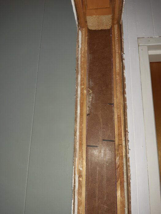 Bilden visar en öppning där en innerdörrs karm tagits bort. Masonit och träskiva är synliga, möjligtvis med tomrum eller reglar bakom.