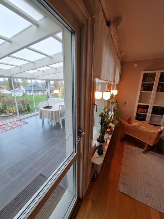 Altandörr inifrån med glipa i övre hörnet; omgivning med glasveranda, bord och stolar utomhus samt inomhusmiljö med bokhylla och krukväxter.