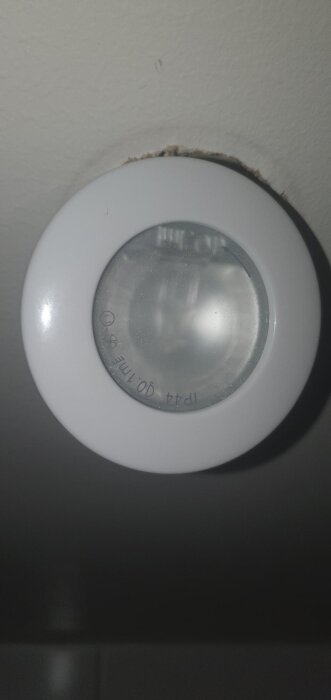 Närbild på en takspotlight med glas och märkning, pil synlig. Den är inpassad i ett hål i taket och omgärdad av en vit ram.