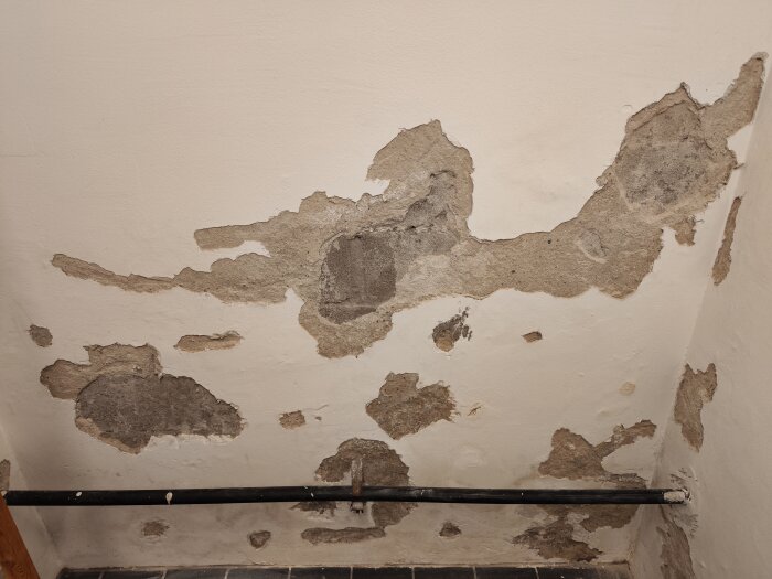 Källarvägg med puts som har släppt, blottar underliggande betong. En svart rörledning löper längs väggen nära golvet.