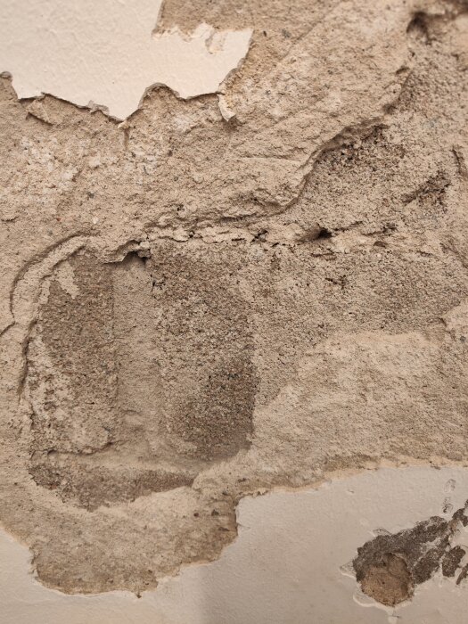 Närbild på en källarvägg med delvis bortknackad puts, exponerade betongblock och flagande färg.