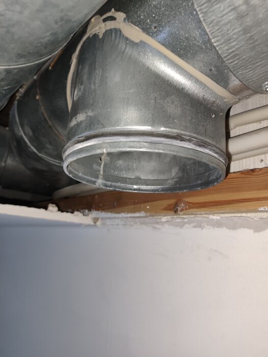 T-rör i metall med ändlock, del av ventilation eller avloppssystem, sett mot en vit vägg och träbjälke.