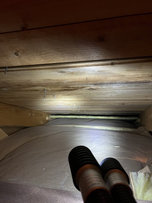 Ett vindsutrymme med virkestak och eventuell fukt, synligt genom inspektion under plastfolie. Ficklampans ljus belyser området.