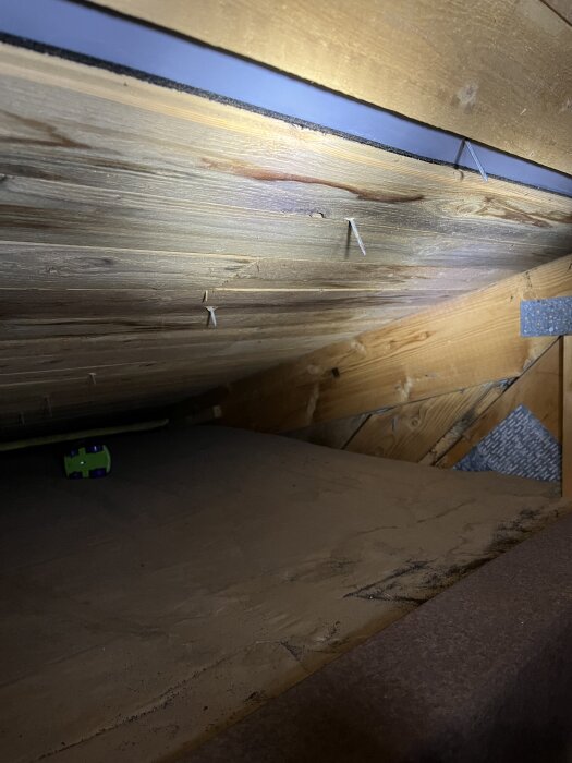 Träbalkar med spikar synliga, en mörk fläck som kan tyda på fukt. En grön leksaksbil syns på den dammiga ytan nedanför.