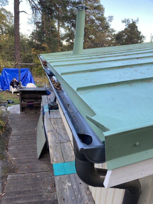 Tak med ny plåt, bytta vindskivor och kvarvarande glipa vid skorstensröret. Skog i bakgrunden och utemöbler täckta av en blå presenning.