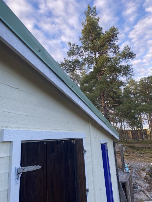 Nymålat hus med bytta vindskivor, fotograferat från sidan. En skog i bakgrunden och molnig himmel ovanför.
