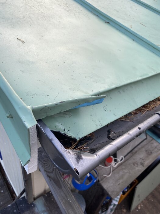 Nära bild på ett grönt plåttak och hängränna med rester av smuts och löv. Detalj av reparationer och hörn av taket syns tydligt.