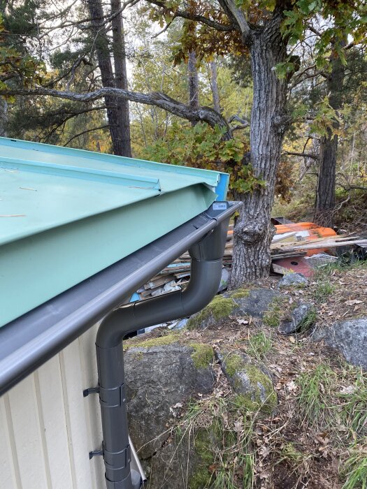 Tak med nymålade vindskivor och nytt stuprör, omgiven av träd och stenar i en skogsmiljö. Problem med glipa och falsning åtgärdade.