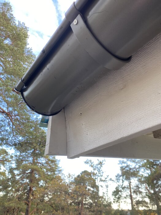 Vitmålad vindskiva och grå stupränna sett underifrån, träd i bakgrunden.