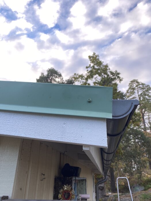 Närbild på takfot med nymålade vindskivor och hängränna, med träd i bakgrunden och del av byggnadsdetalj.