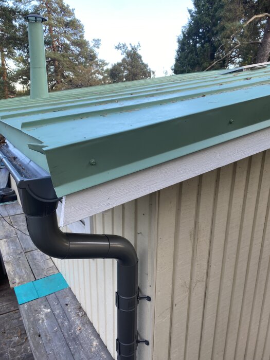 Grönt plåttak med nya vindskivor, synlig regnvattensrör och skorstensrör, omgiven av träd. Taket nyligen renoverat och målat.