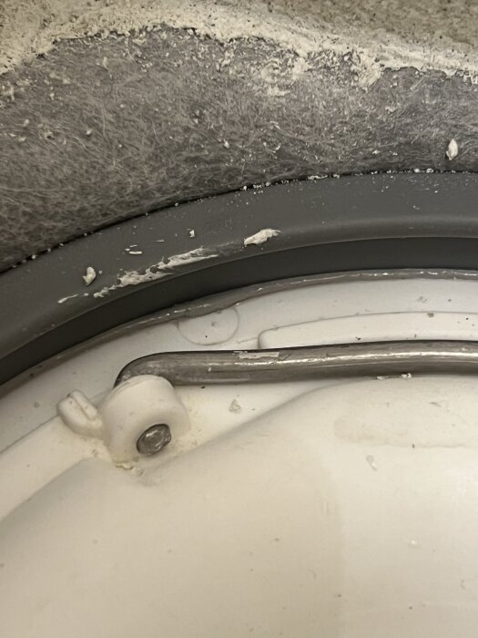 Metallklämma och skruv på en vit yta med fogmassa och damm i bakgrunden.