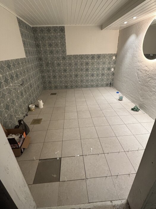 Badrum under renovering med nylagda grå klinkersplattor, vägg med mönstrade kakelplattor, kartong och rördelar synliga.