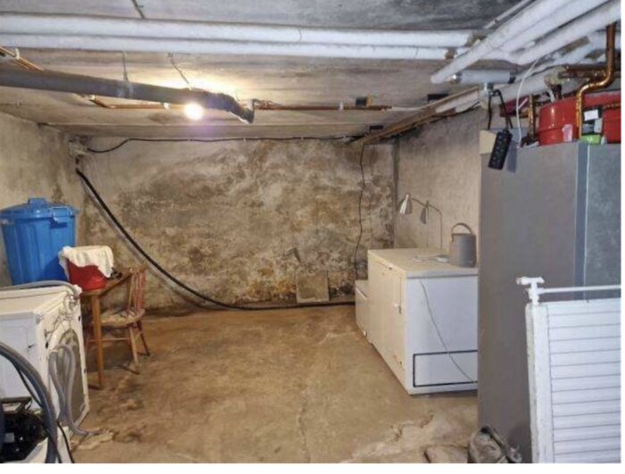 Fuktig källare med missfärgade väggar, tvättmaskin, blå tunna och rör i taket. Diskussion om målning och dränering för att minska fukt.