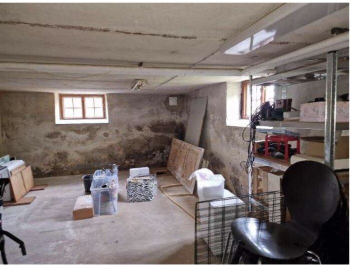 Fuktig källare med missfärgade väggar, lagerhyllor och kartonger. Fönster med ljusinsläpp, cementgolv och lagrade föremål syns.