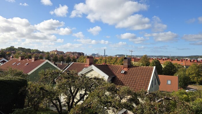 Taklandskap med röda tegeltak och omgivande grönska under en delvis molnig himmel. Avlägsna byggnader syns i bakgrunden.