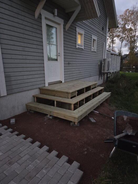 Nysnickrade trappsteg av trä vid grovköksentrén, omgivna av grått tegel och brun jord. Huset har grå träfasad med en vit dörr och fönster.