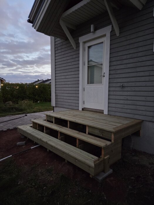 Trätrappa vid ytterdörr, nyligen byggd av träplankor, mot ett gråmålat hus.