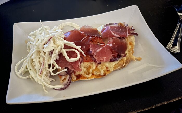 Pinsa med prosciutto, rödlök och mozzarella på tallrik, serverad med coleslaw.