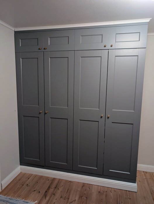 Grå PAX-garderob med egengjorda överskåp av mdf, guldfärgade knoppar, och taklister, stående på trägolv mot ljus vägg.