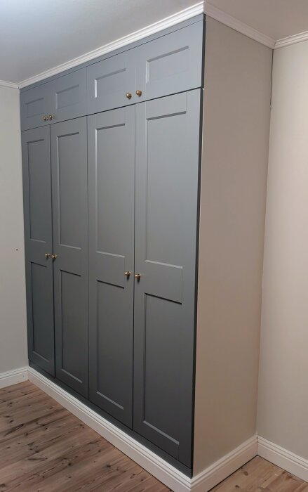 Grå PAX-garderober med egna överskåp monterade mot en ljus vägg, med mässingknoppar och vit sockel samt taklist.