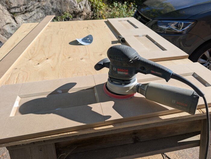 Slipmaskin på en arbetsyta med mdf-paneler för byggande av överskåp, i solen vid en bil och bergvägg i bakgrunden.