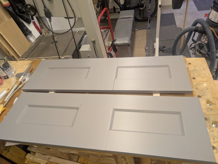 Gråa MDF-dörrar med spegelpaneler liggande på en arbetsbänk i verkstadsmiljö, omgivna av verktyg och maskiner.