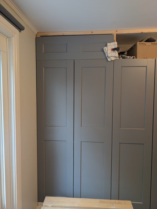 Gråa garderober med hemmagjorda överskåp av mdf, integrerade i rummet vid taket, med pågående arbete med tyg och verktyg ovanpå.