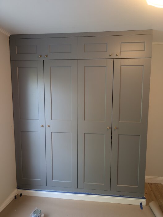 Grå PAX-garderob med egenbyggda överskåp, mässingsknoppar och taklister, placerad i ett rum med ljusa väggar och trägolv.