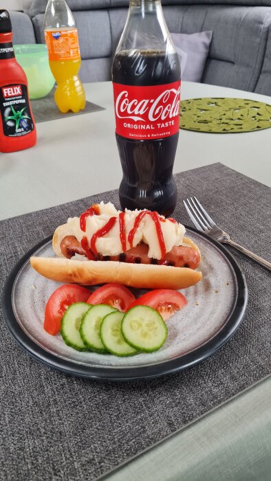 Korv med potatismos och ketchup serverad på tallrik med tomat och gurka. Flaskor med Coca-Cola och Fanta i bakgrunden.