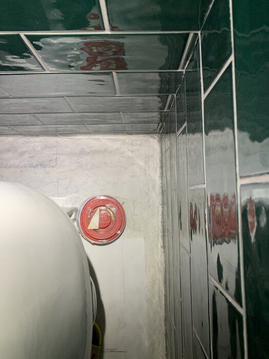 Badrumshörn med gröna kakelplattor, avloppsanslutning nära högervägg. Toalett bakom kameran, röd lock på avloppsanslutningen synlig.