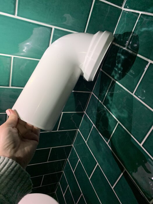 Person håller ett böjt vitt rör i badrum, nära grön kaklad vägg. Rör används som alternativ lösning för toalettens avloppsplacering.